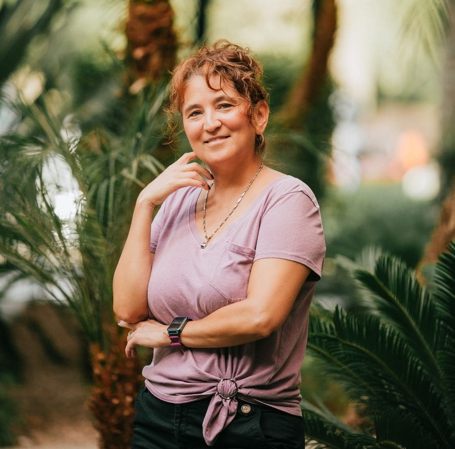 Smiling woman in a pink tied-front t-shirt standing among lush tropical plants outdoors.